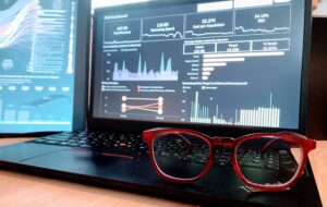 Red framed eye glasses in focus on the bottom right foreground, placed on a Thinkpad laptop displaying a dark themed analytics dashboard