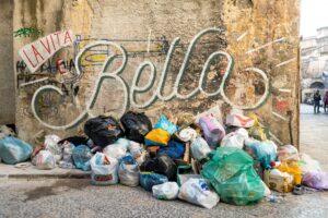 Pile of garbage bags in front of a dirty yellow wall boasting a large graffiti stating "La Vita E Bella"