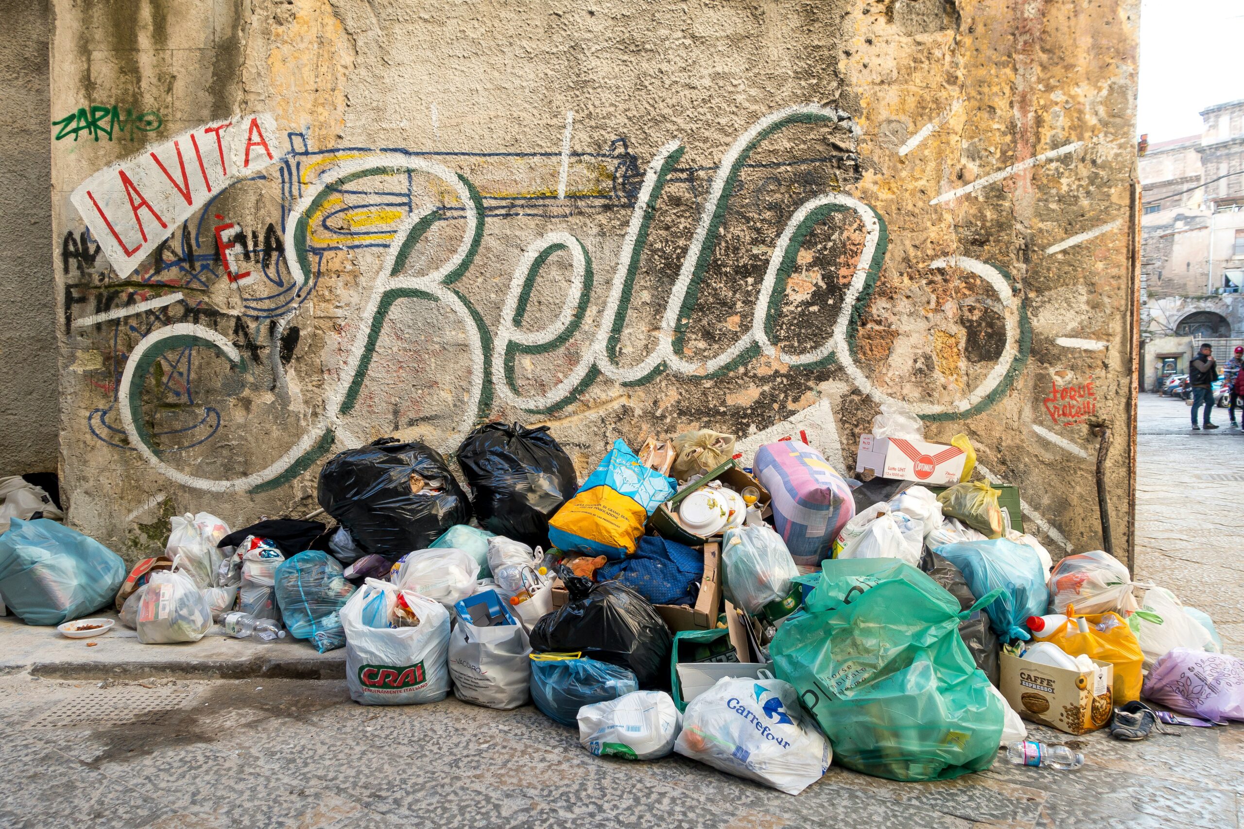 Pile of garbage bags in front of a dirty yellow wall boasting a large graffiti stating "La Vita E Bella"