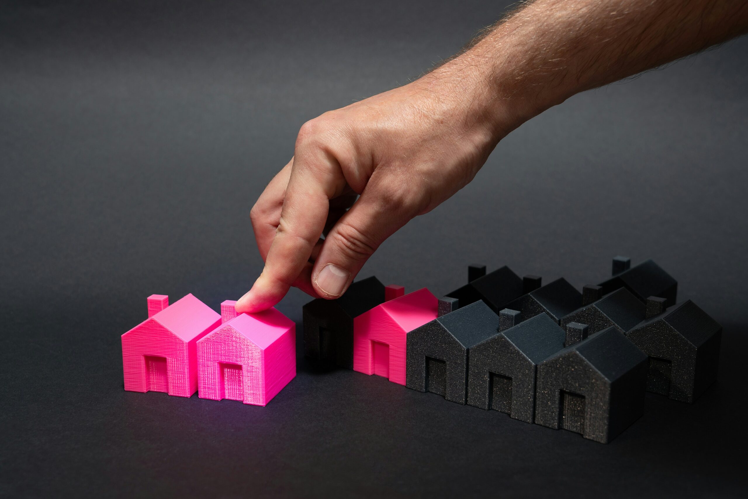 A hand reaching for a little pink Monopoly style house with other similar black and pink houses around on a black background