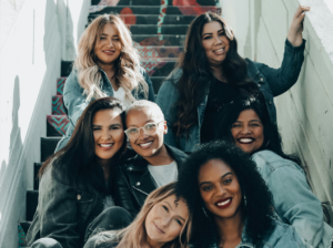 7 women of different ethnicities in casual clothing sitting on a staircase smiling